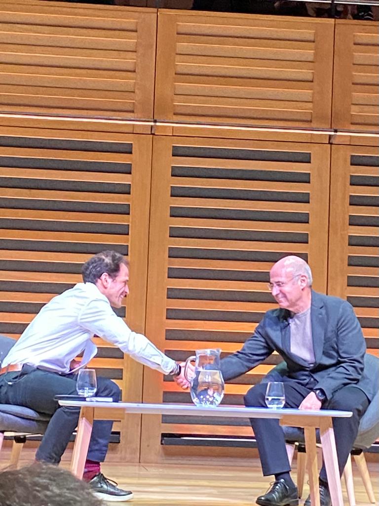 Daniel Ross and Bill Browder seated on stage, shaking hands across a low table.